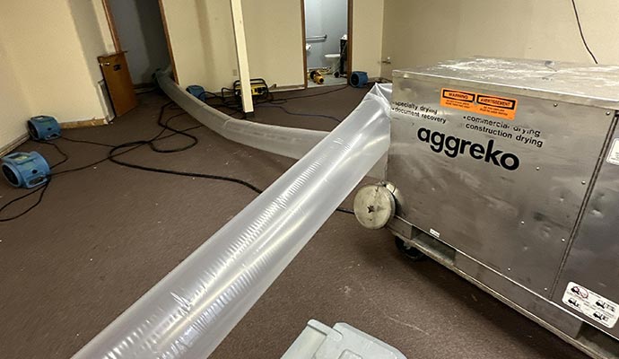 A large Aggreko industrial desiccant dehumidifier with plastic ducting and air movers drying out a water-damaged room