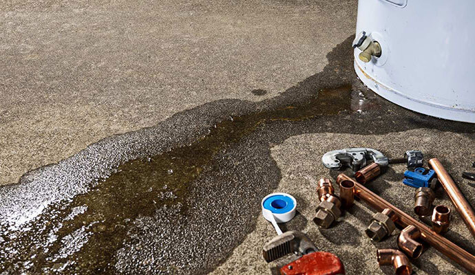 Leaking Water Heater with Plumbing Tools A leaking water heater with water pooling on the floor, surrounded by plumbing tools and fittings.